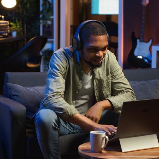 Focused black guy teleworking on laptop in a living room feels happy with task completion, achieving success. Remote worker balancing comfort and work efficiency in a flexible workspace. Camera B.